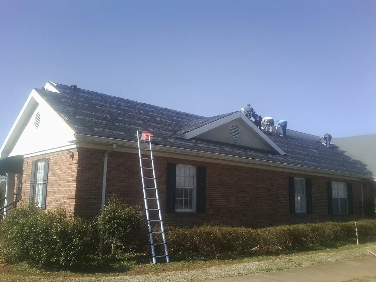 Crew tearing shingles off a brick ranch roof with a ladder set — Emergency Roof Repair work by our crew in Mondamin