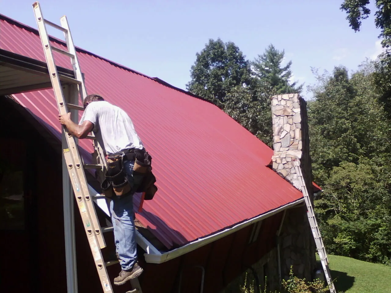 Roofer on a ladder beside a red metal roof and stone chimney — Soffit & Fascia Repair by Roofing Brothers in Mondamin